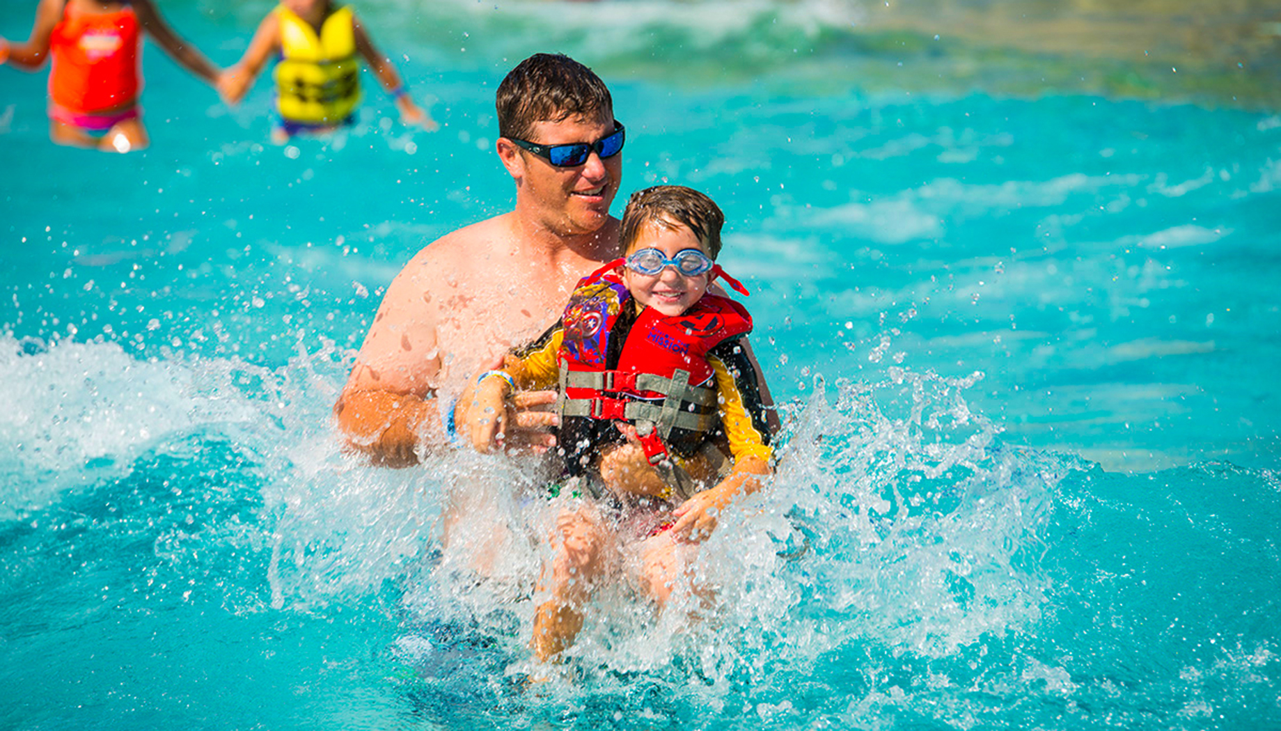 An adult and child wearing life vests and goggles play in a pool with splashing water. Other people are visible in the background.